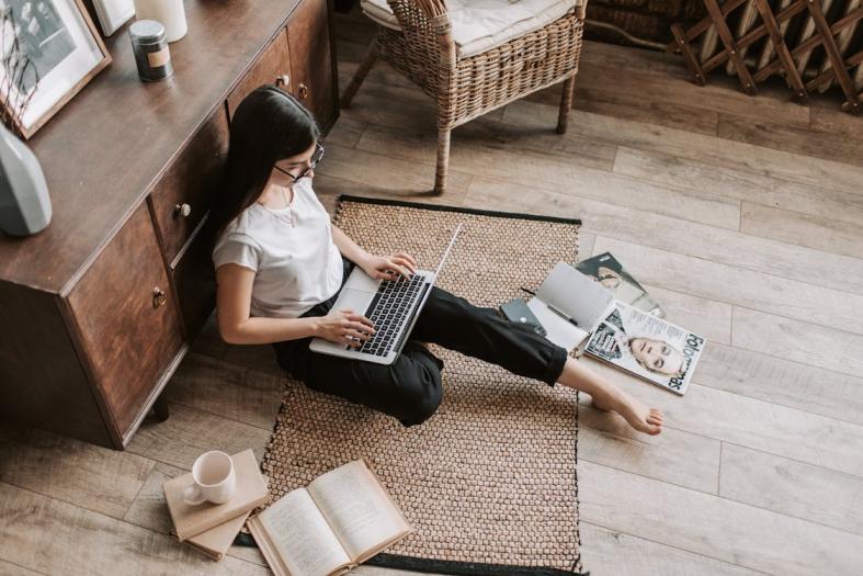 A woman sitting on the floor, focused on her laptop, with a relaxed posture and a cozy environment around her.