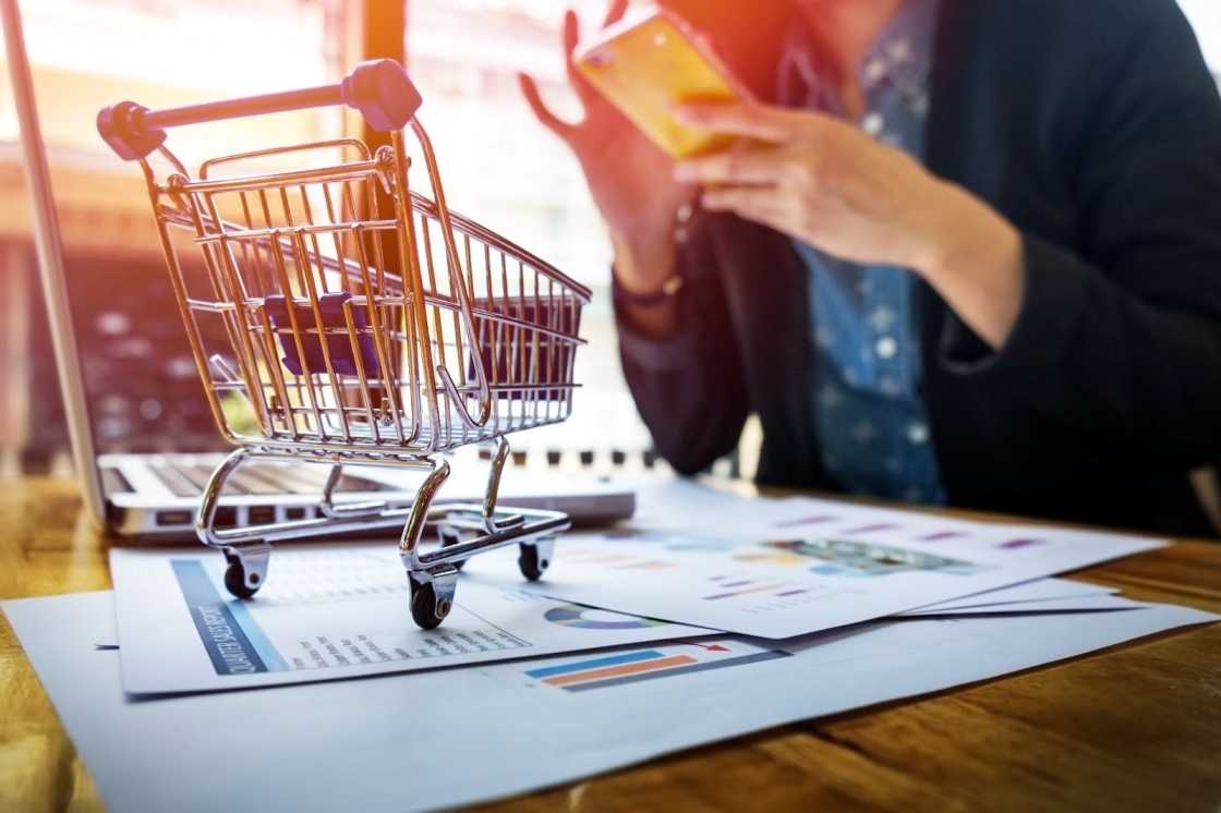 A woman uses her phone at a desk, with a shopping cart nearby